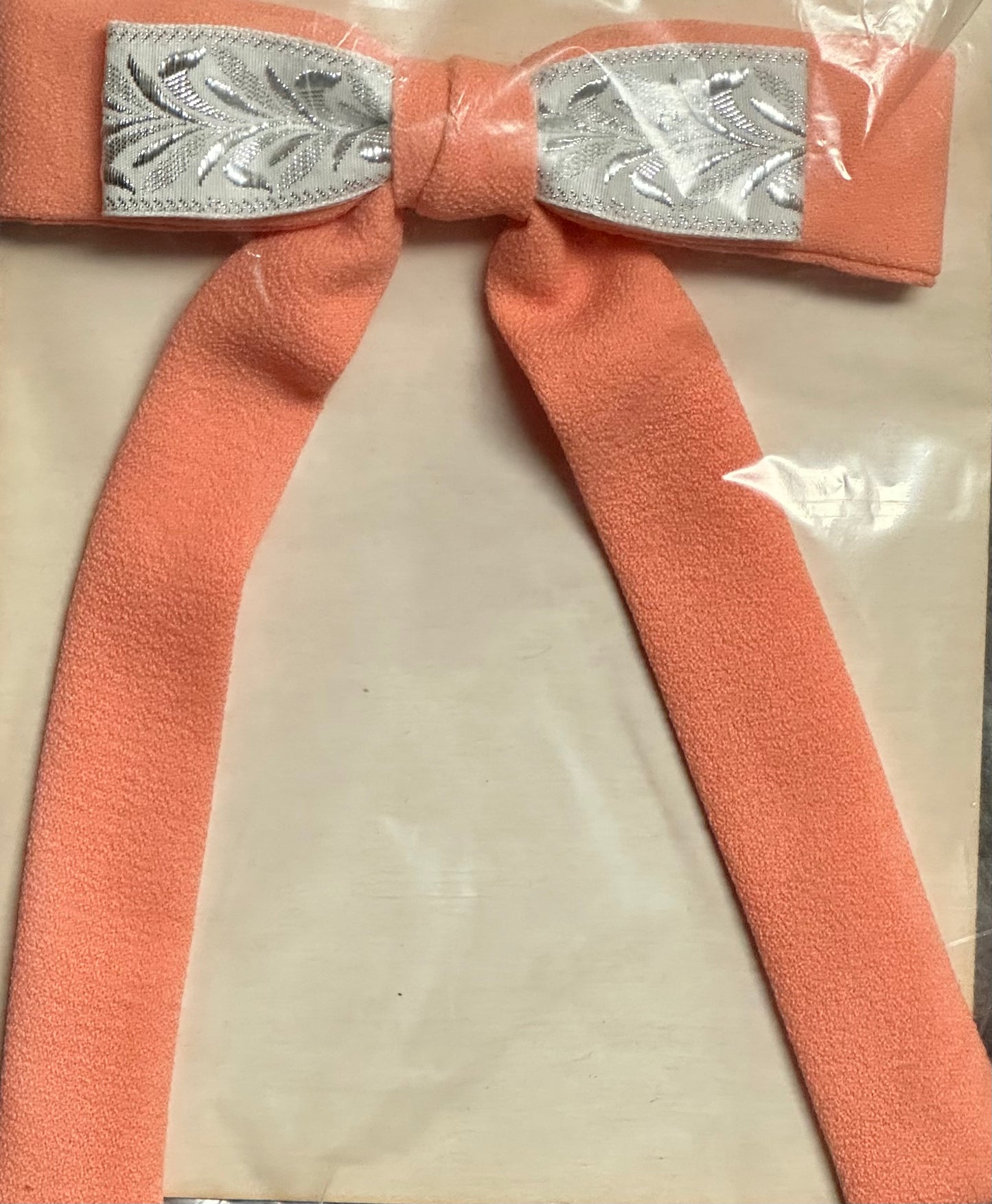 The Colonel Ties by Square Up Fashions features a peach-colored bow with silver leaf-patterned ribbon on a plastic-wrapped background, evoking the charm of a southern bow tie.