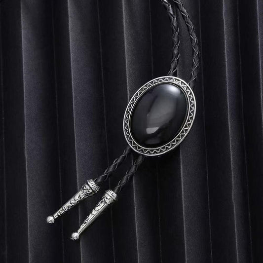 The Western Express Black Stone Bolo Tie features a braided leather cord, silver tips, and an oval black stone clasp—an elegant western accessory displayed on a pleated black background.