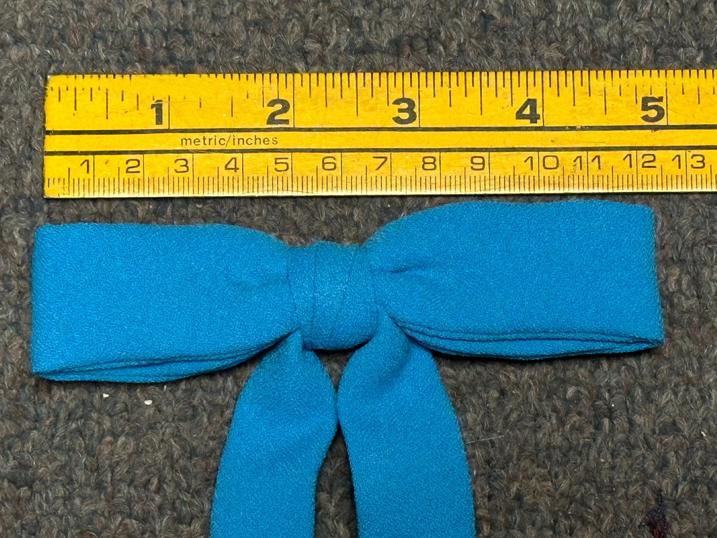 A Colonel Tie by Square Up Fashions, in stylish blue fabric, is elegantly positioned below a yellow ruler that features both metric and inches measurements, resting on a gray surface.