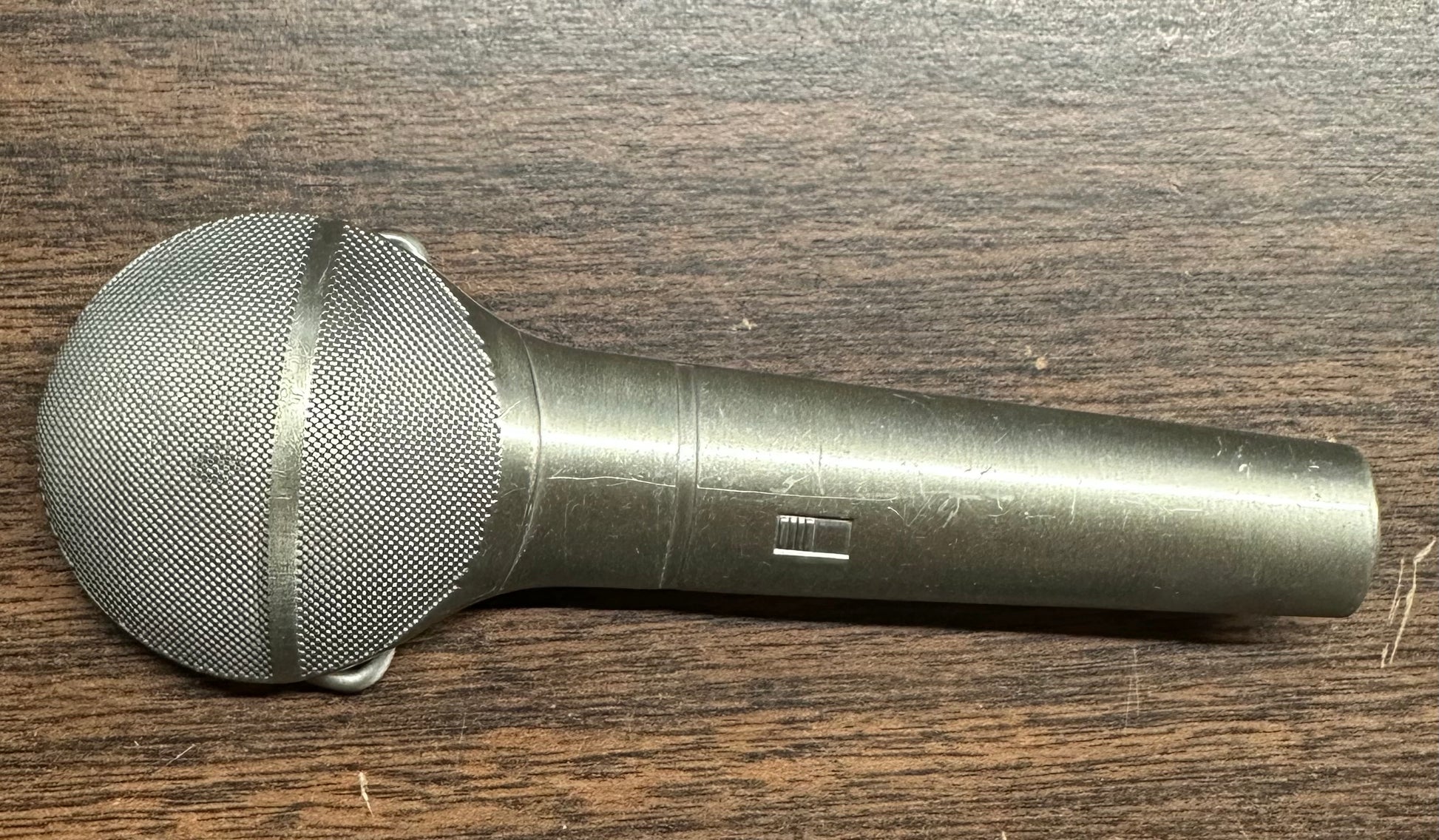 The Square Up Fashions SD Callers’ Belt Buckle, designed to resemble a silver handheld microphone with a mesh grille, rests on a dark wooden surface.