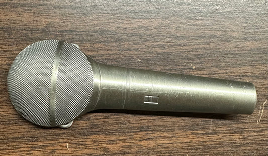 The Square Up Fashions SD Callers’ Belt Buckle, designed to resemble a silver handheld microphone with a mesh grille, rests on a dark wooden surface.