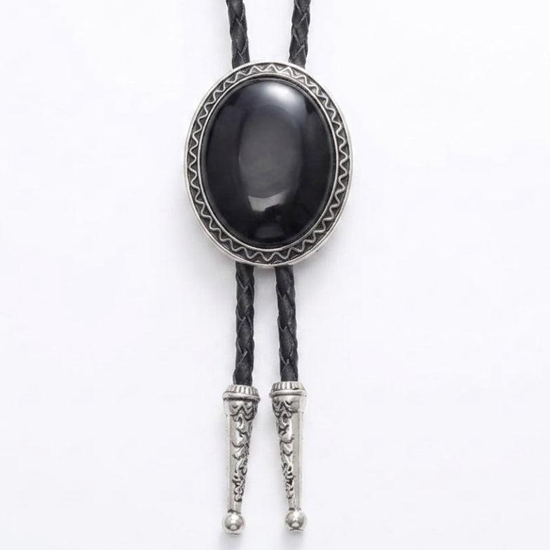 A black bolo tie necklace featuring a large oval black onyx cabochon stone surrounded by a silver-toned geometric bezel, with braided black cords and silver-toned decorative tip connectors.
