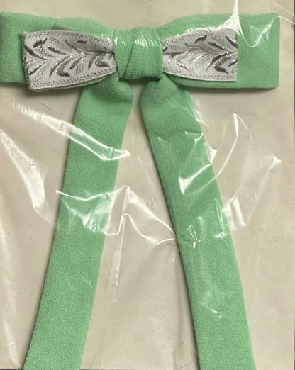 Introducing the Colonel Ties by Square Up Fashions: a charming southern bow tie showcasing a green bow with long tails, elegantly accented by a silver patterned center, and neatly wrapped in clear plastic packaging.