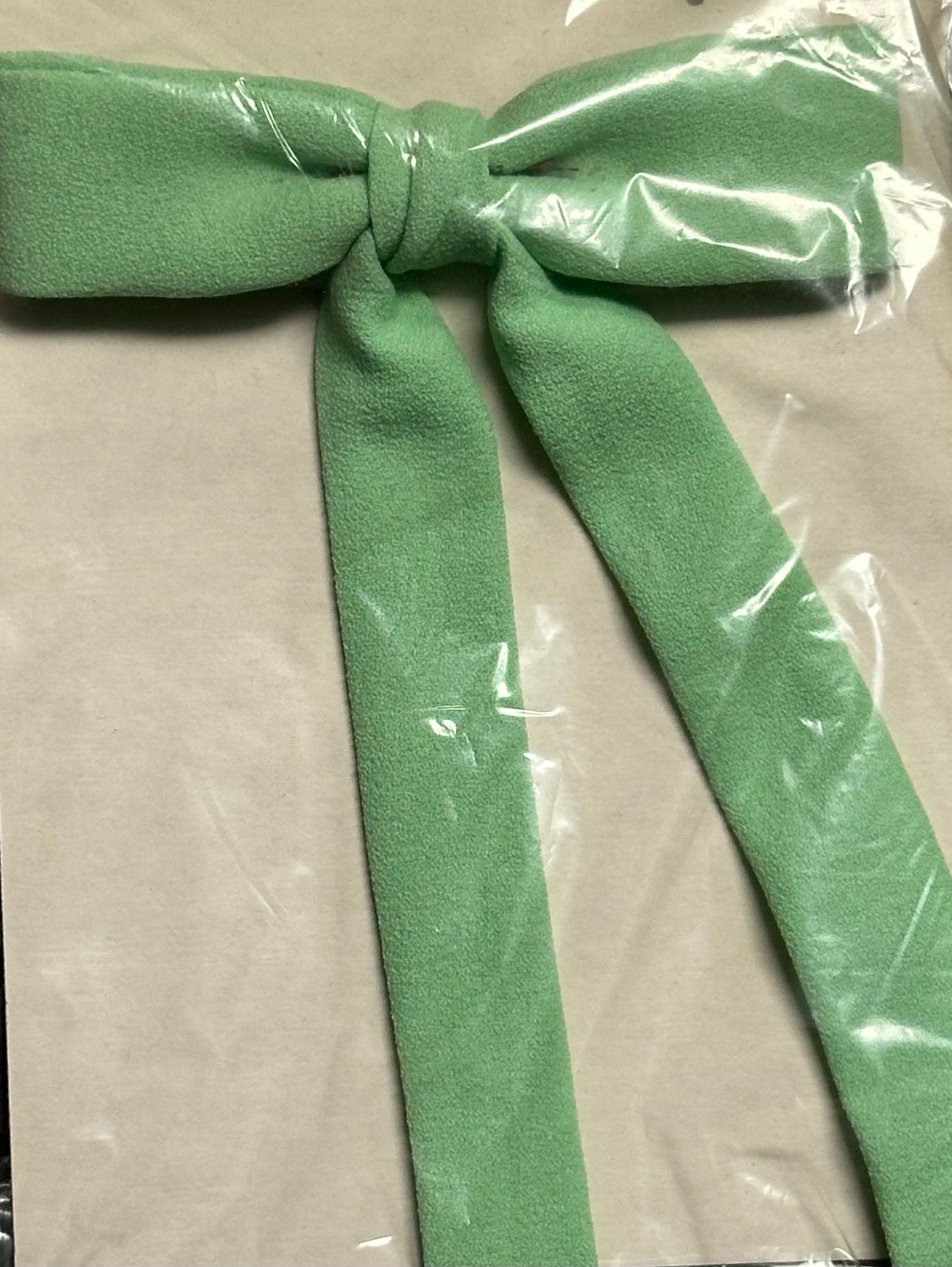 A stylish green Colonel Tie by Square Up Fashions, reminiscent of a southern bow tie, is elegantly tied against a beige background and wrapped in clear plastic.