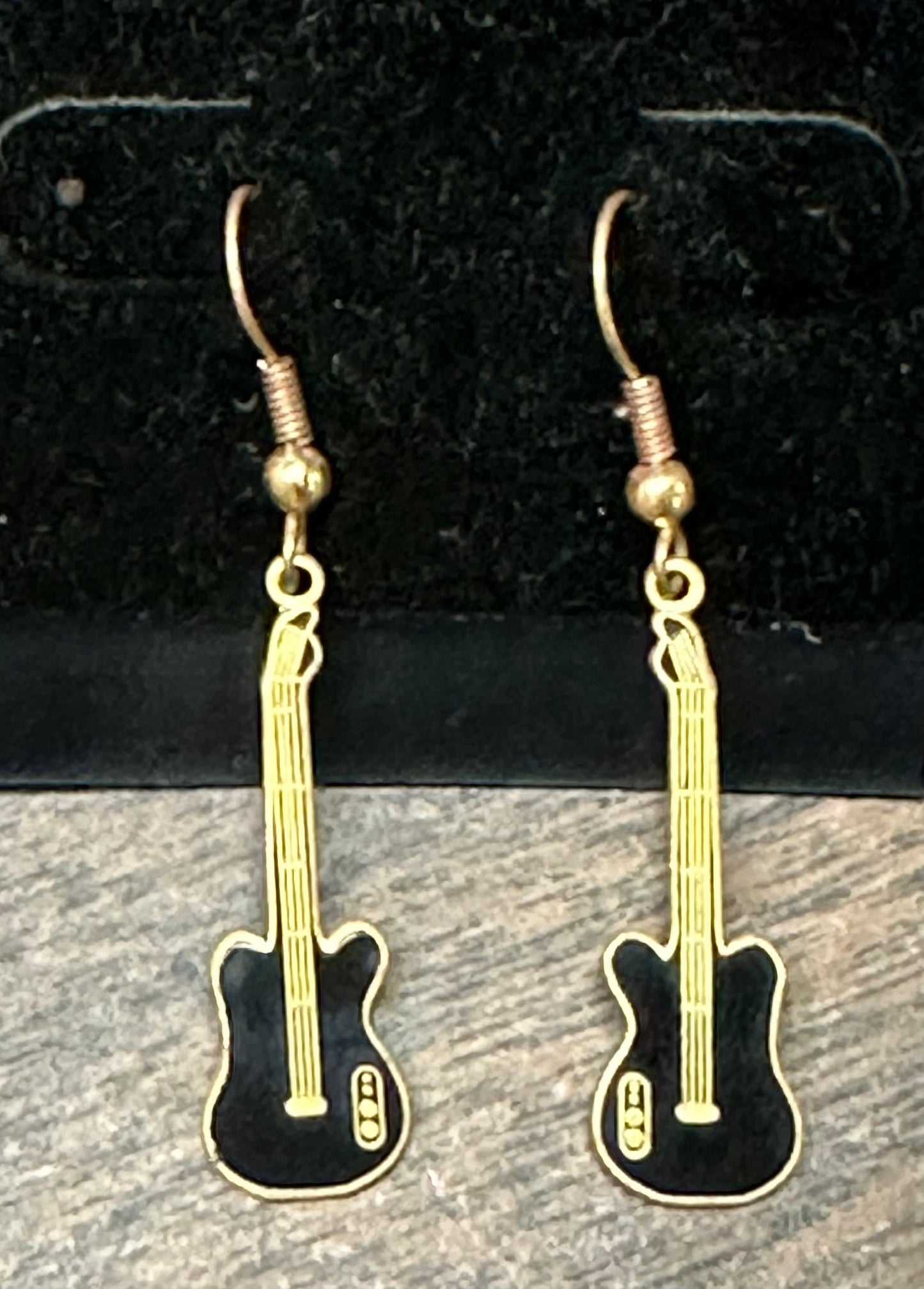 These Guitar Dangle Earrings by Square Up Fashions feature a design resembling black electric guitars with golden strings and hooks, perfectly capturing the essence of country guitars against a dark background.