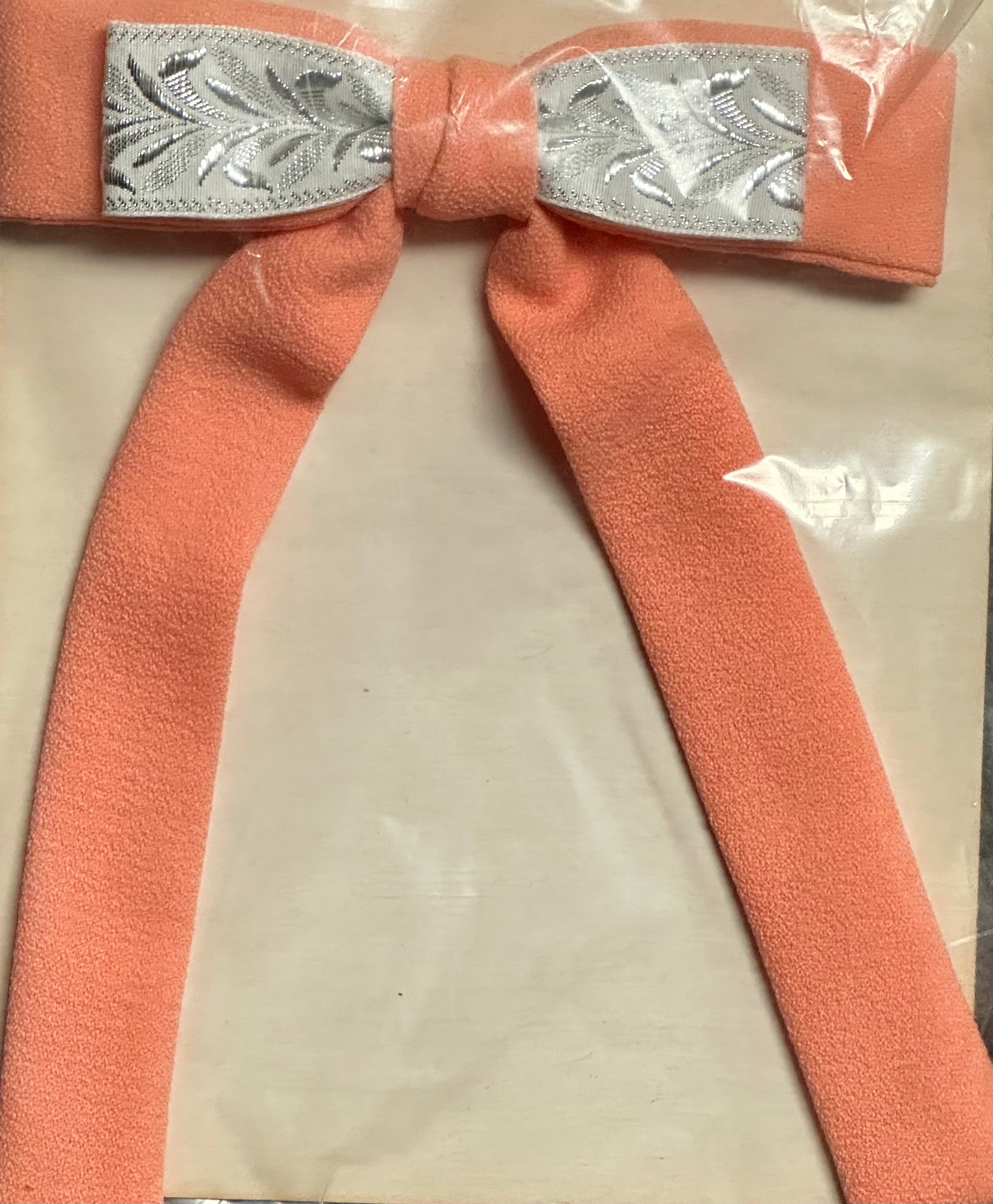 The Colonel Ties by Square Up Fashions features a peach-colored bow with silver leaf-patterned ribbon on a plastic-wrapped background, evoking the charm of a southern bow tie.