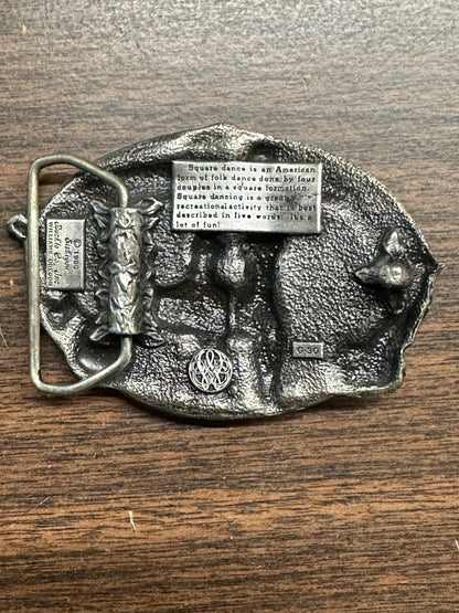 Square Up Fashions' Square Dance Buckle w/ instruments features an engraved definition, relief dancer, decorative patterns, and "DO SI DO." The buckle is shown resting on a wooden surface.