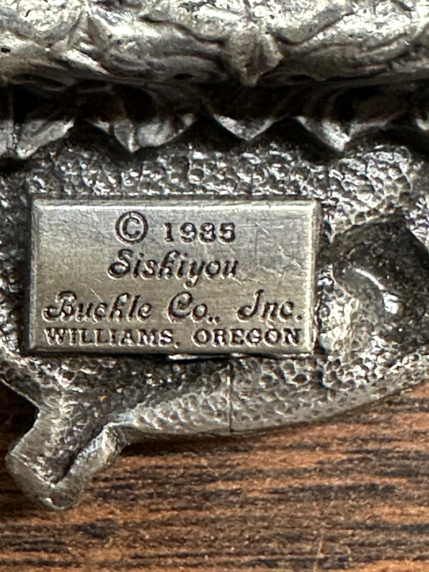 Close-up of a metal plaque reading "Square Dance Buckle w/ instruments" by Square Up Fashions, embossed on a textured surface, reminiscent of detailing found on vintage Anaheim belt buckles.