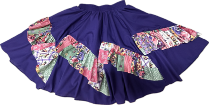 A colorful Diagonal Pinwheel Square Dance Skirt with vibrant print ruffle and floral designs displayed on a contrasting solid purple background by Square Up Fashions.