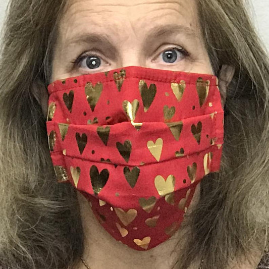 A woman wearing a Square Up Fashions red face mask with gold hearts on it. The face mask is washable and reusable, and features an adjustable nose wire for added comfort. It also provides 2 layers of